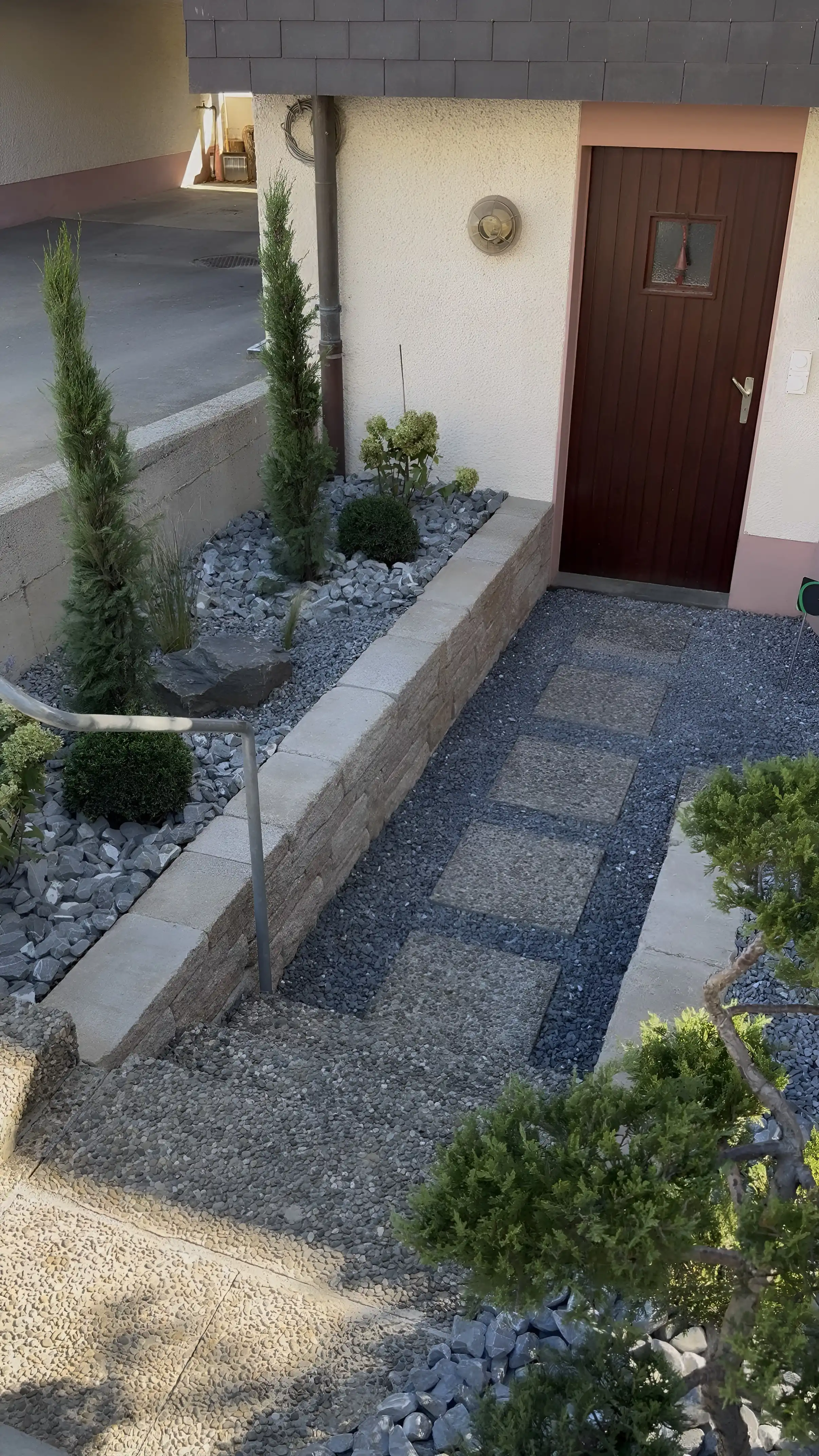 Allée en pas japonais jusqu'à une porte d'entrée : dalles dans le gravier gris, muret en pierre, conifères en colonne